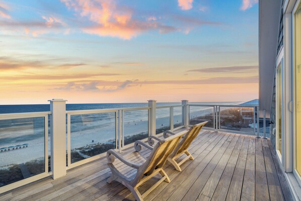 A wooden deck with two lounge chairs overlooking a beach at sunset.