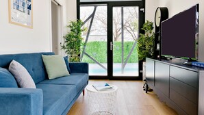 A modern living room with a blue sofa, a white coffee table, a flat-screen TV on a cabinet, and a view of a green hedge and trees through a glass door. - Modern Villa Barcolana with a swimming pool (Baderna)