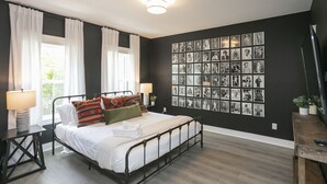 Un dormitorio con estructura de cama metálica, ropa de cama blanca, una pared de fotografías en blanco y negro, y una mesita de noche de madera con lámpara.