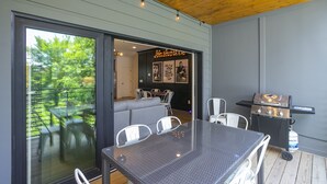 Un área de comedor al aire libre con mesa y sillas de vidrio, una parrilla y vista del jardín a través de la puerta de cristal.