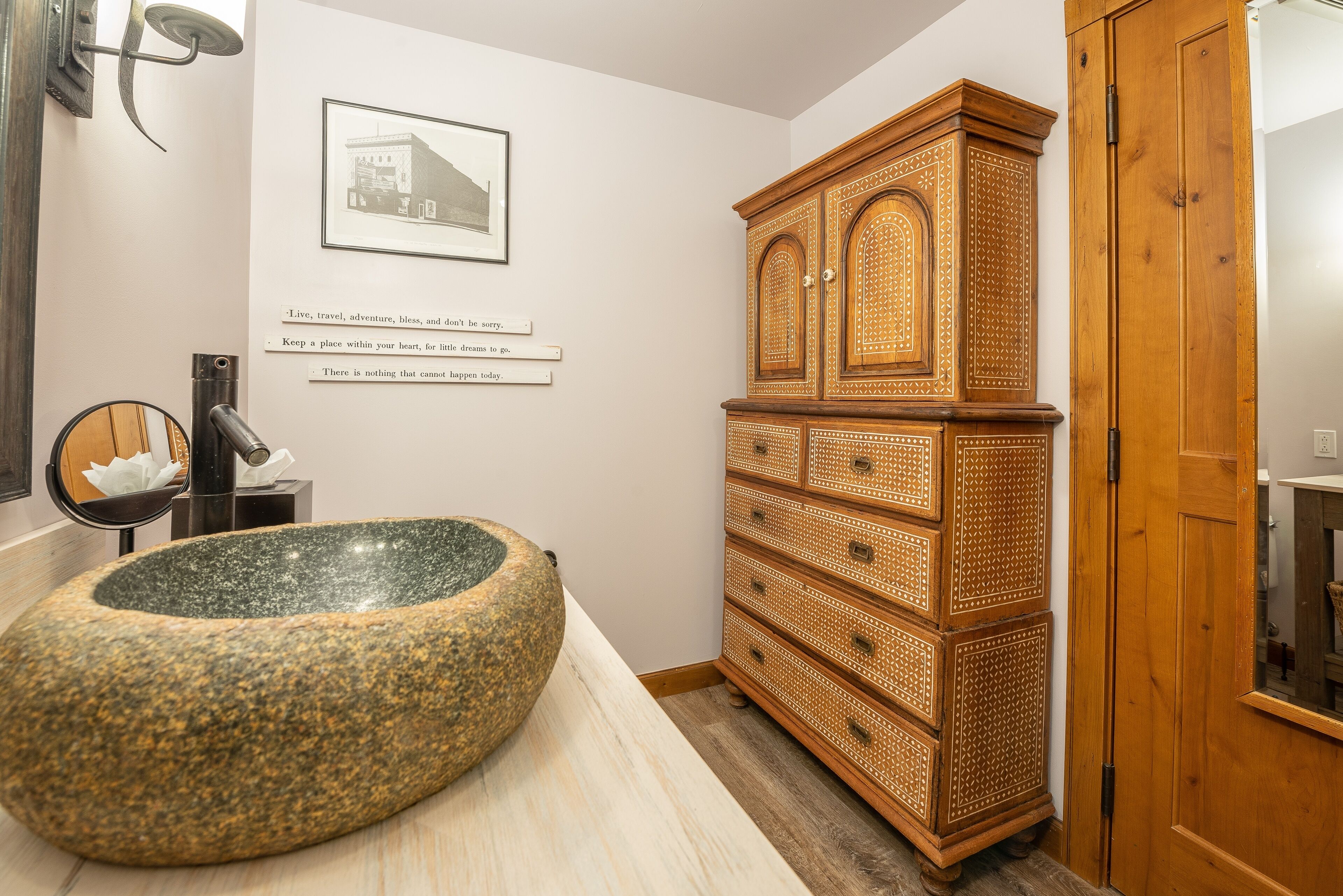 A bathroom with a large stone basin, a wooden cabinet with drawers, a framed picture on the wall, and a mirror with a towel.