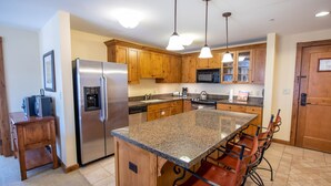 A kitchen with wooden cabinets, a stainless steel refrigerator, granite countertops, and bar stools. - Spacious 3 Bedroom Condo with Balcony in Mountaineer Square 3 Condo - No Cleaning Fee! by RedAwning (Crested Butte)