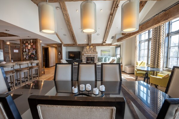 A spacious living room with a large glass table, multiple seating options, a stone fireplace, and a bar area with stools.