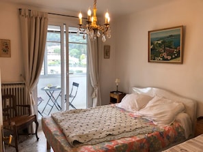 A hotel room with a bed, a chair, a table, a radiator, a chandelier, and a painting on the wall.