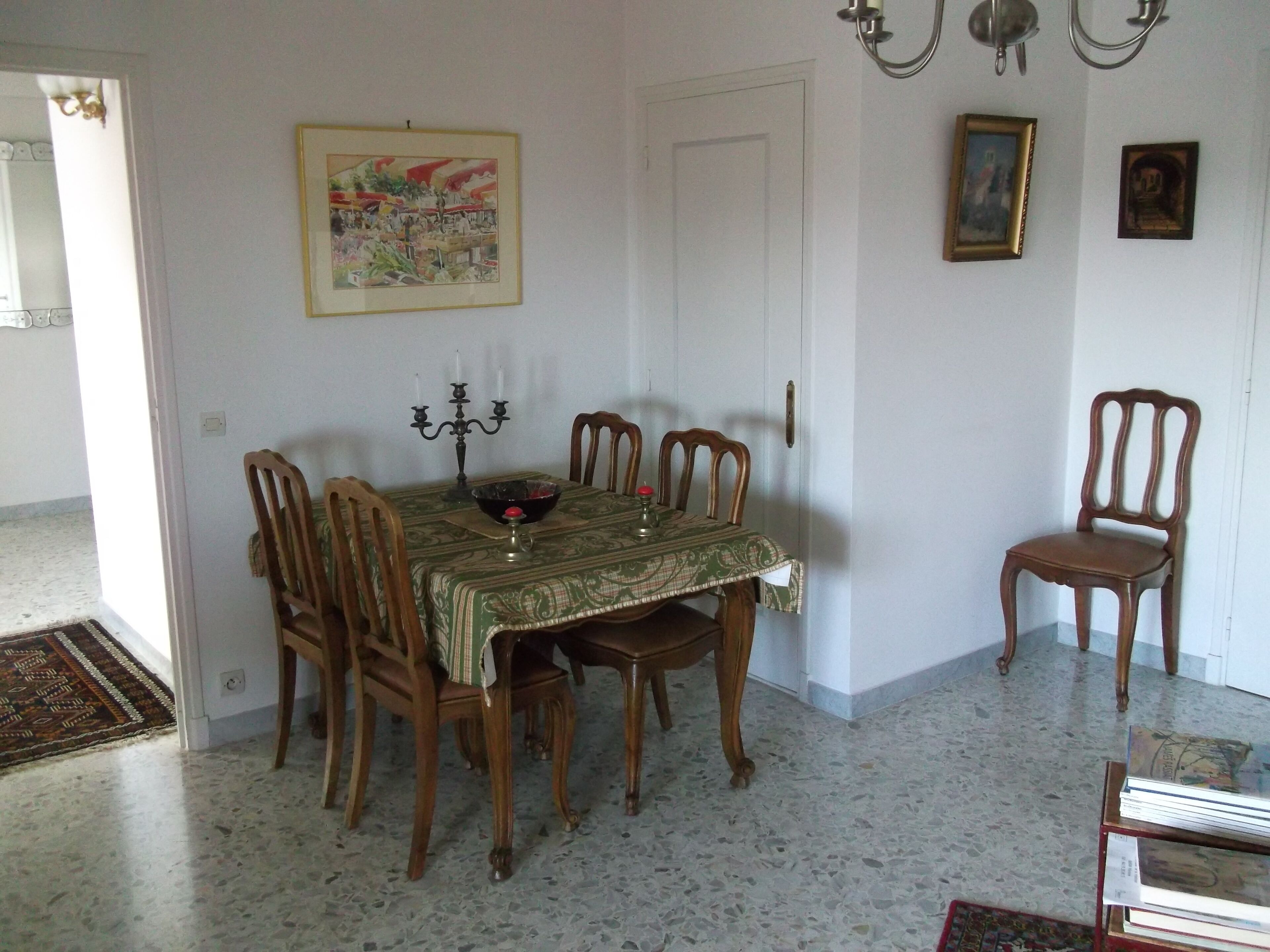 Un comedor con una mesa dispuesta para cuatro personas, rodeada de sillas de madera. Un candelabro cuelga sobre la mesa, y hay un cuadro enmarcado en la pared.