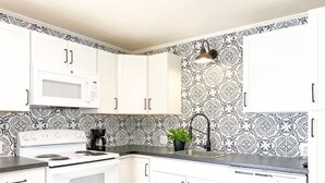 Une cuisine avec des meubles blancs, un four à micro-ondes, une cuisinière, un évier et un crédence décorée.