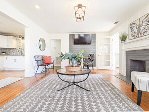 A living room with a round coffee table, a fireplace, a television, and a kitchen in the background. - The Nook on Main ~ Walk to restaurants and shops! (Mountain View)