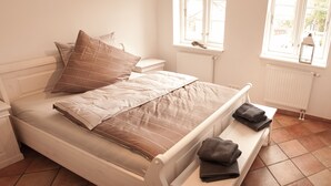 A neatly made bed with a striped blanket and pillows, a bedside table with a lamp, and a window with a view of the outside. - Das Altenteil (Stoltebuell)