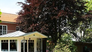 A covered patio with a brick wall, a lawn, and a large tree with red leaves. - Das Backhaus 2 (Stoltebuell)