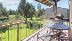 Een balkon met uitzicht op een gazon en bomen, een barbecue en een tafel met stoelen.