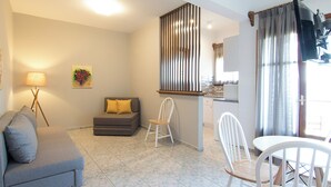 A modern living room with a grey sofa, a small dining table with white chairs, a kitchen area, and a large window with curtains. - Olga Villa (Skiathos)
