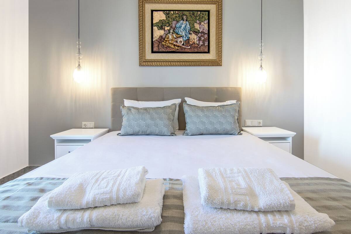 Una cama bien hecha con sábanas blancas y dos almohadas, una mesita de noche con una lámpara y un cuadro en la pared.
