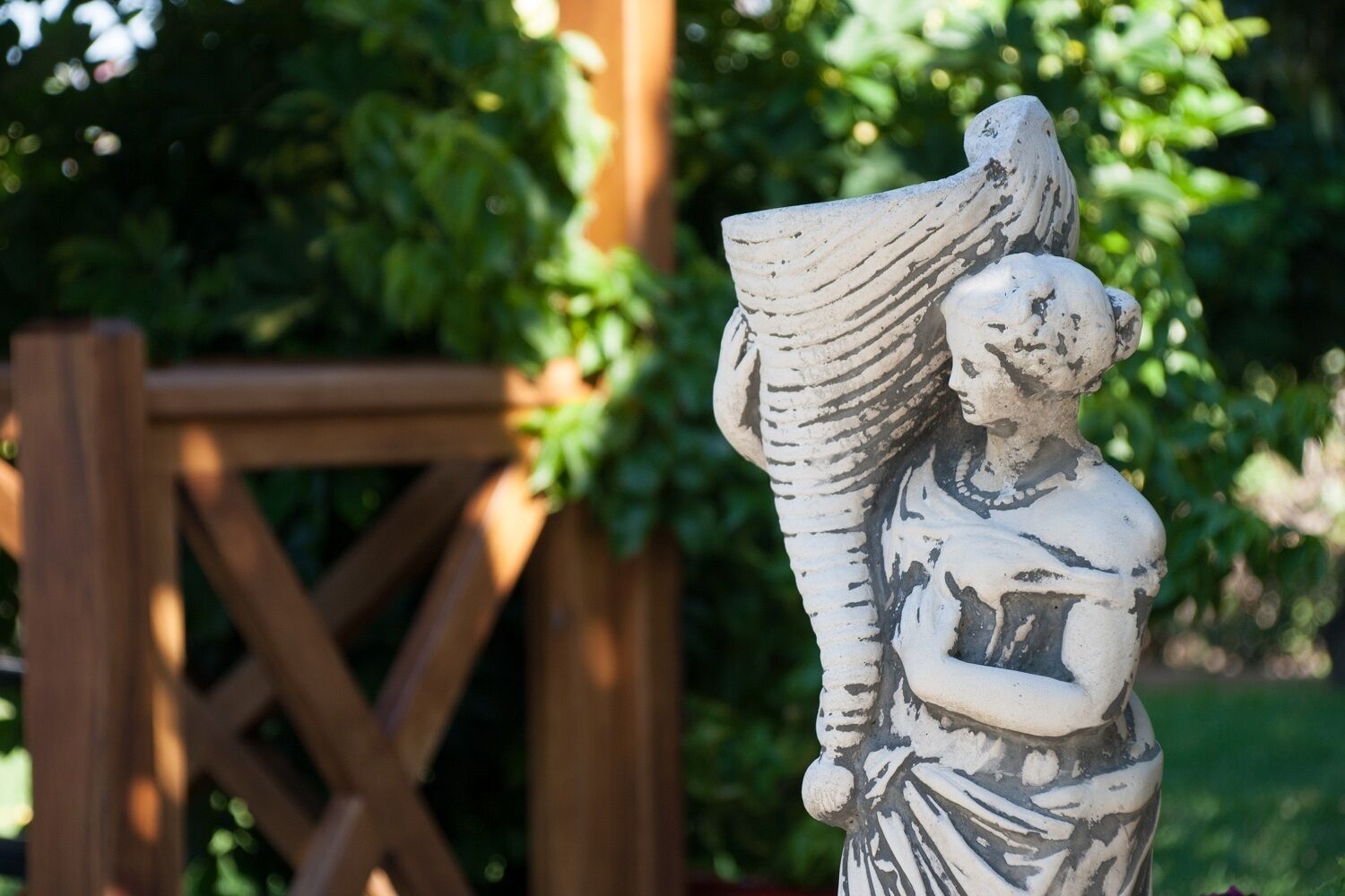 A statue of a figure holding a large object, set against a wooden fence and greenery.