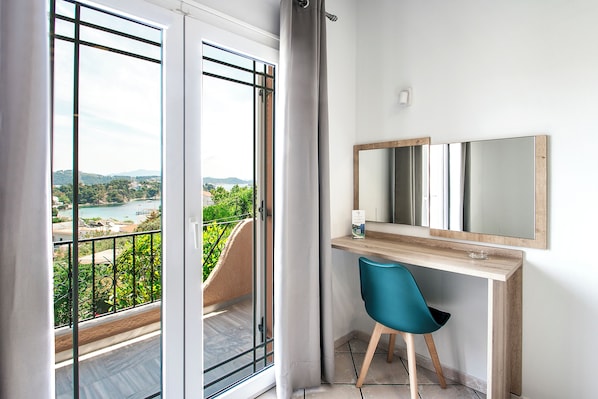 Une chambre avec un balcon, un bureau avec une chaise, un grand miroir et une vue sur une étendue d’eau et sur la verdure.