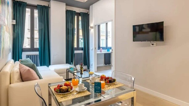 A modern living room with a sofa, a glass coffee table with breakfast items, and a TV mounted on the wall. - Sweett | Duomo 2A - Two Bedroom Apartment, Sleeps 4 (Milano)