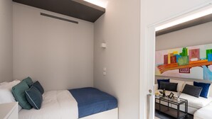 Une petite chambre à coucher moderne, avec un lit, un mur blanc, une bouche d’aération au plafond et une porte donnant sur un espace de vie comprenant un canapé et un tableau coloré.