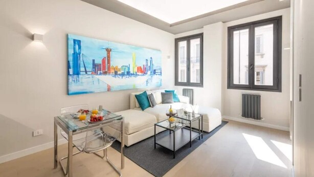 Een moderne woonkamer met een witte bank, een glazen salontafel en een schilderij van een stadsgezicht.