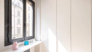 Een kamer met een raam, een radiator, een roze vaas en een blauw bord op een witte plank.