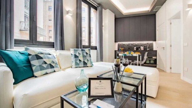 A modern living room with a white sofa, patterned cushions, a glass coffee table with a certificate, and a kitchen area in the background.
