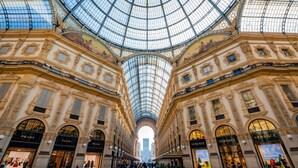 A grand, ornate building with a glass dome ceiling and multiple storefronts, including Prada and Versace.