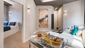 A modern hotel room with a glass dining table set for breakfast, a bed with white bedding, and a large abstract painting on the wall.