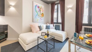 A modern hotel room with a sofa, a glass coffee table, a dining table with breakfast items, a painting on the wall, and a view of the street through large windows.