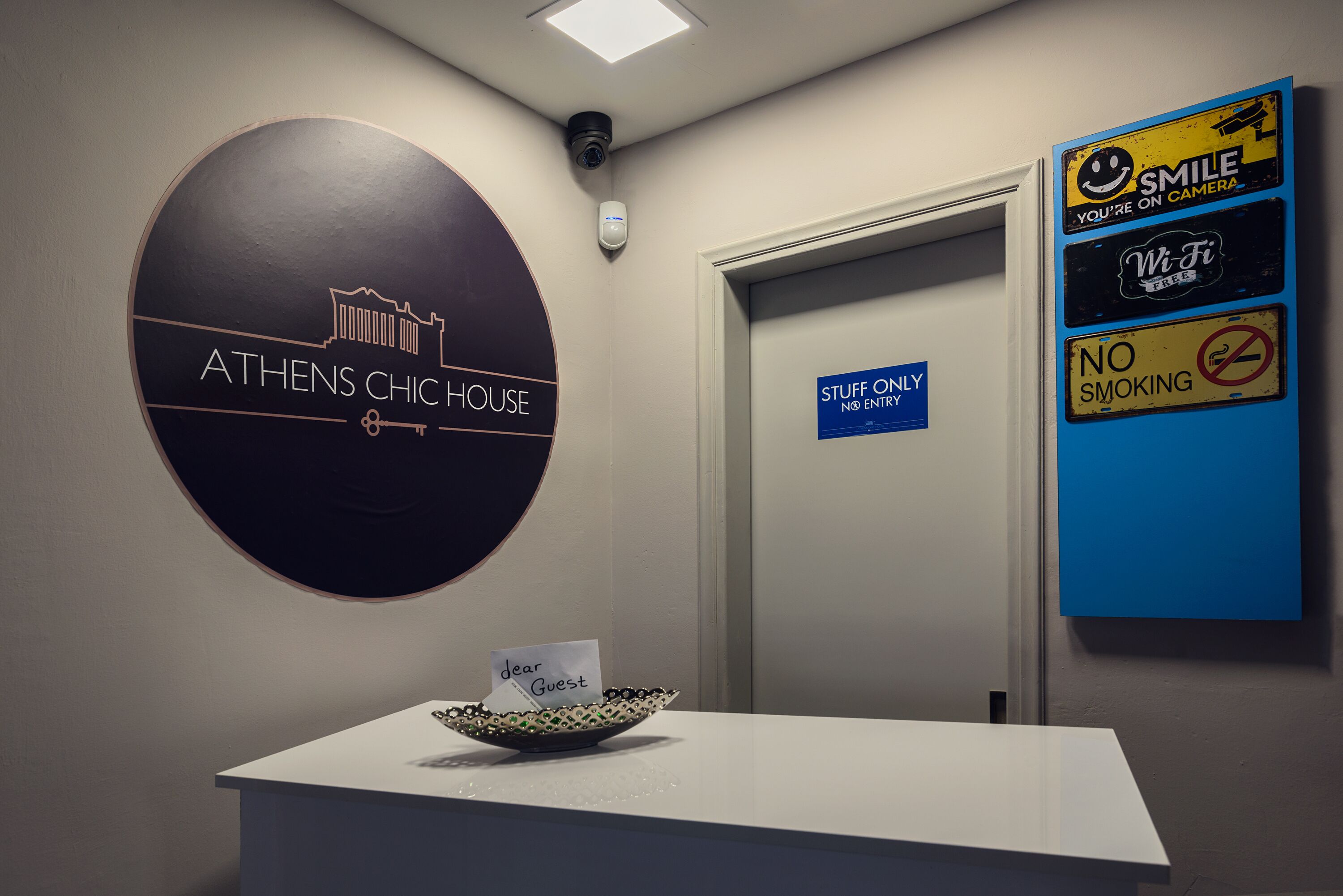 Un comptoir d’accueil avec une pancarte indiquant « ATHENS CHIC HOUSE » et une autre pancarte indiquant « RÉSERVÉ AU PERSONNEL, L’ACCÈS EST INTERDIT ».
