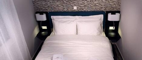 A hotel room with a stone wall, a bed with white linens, two bedside lamps, and two folded towels on the bed.