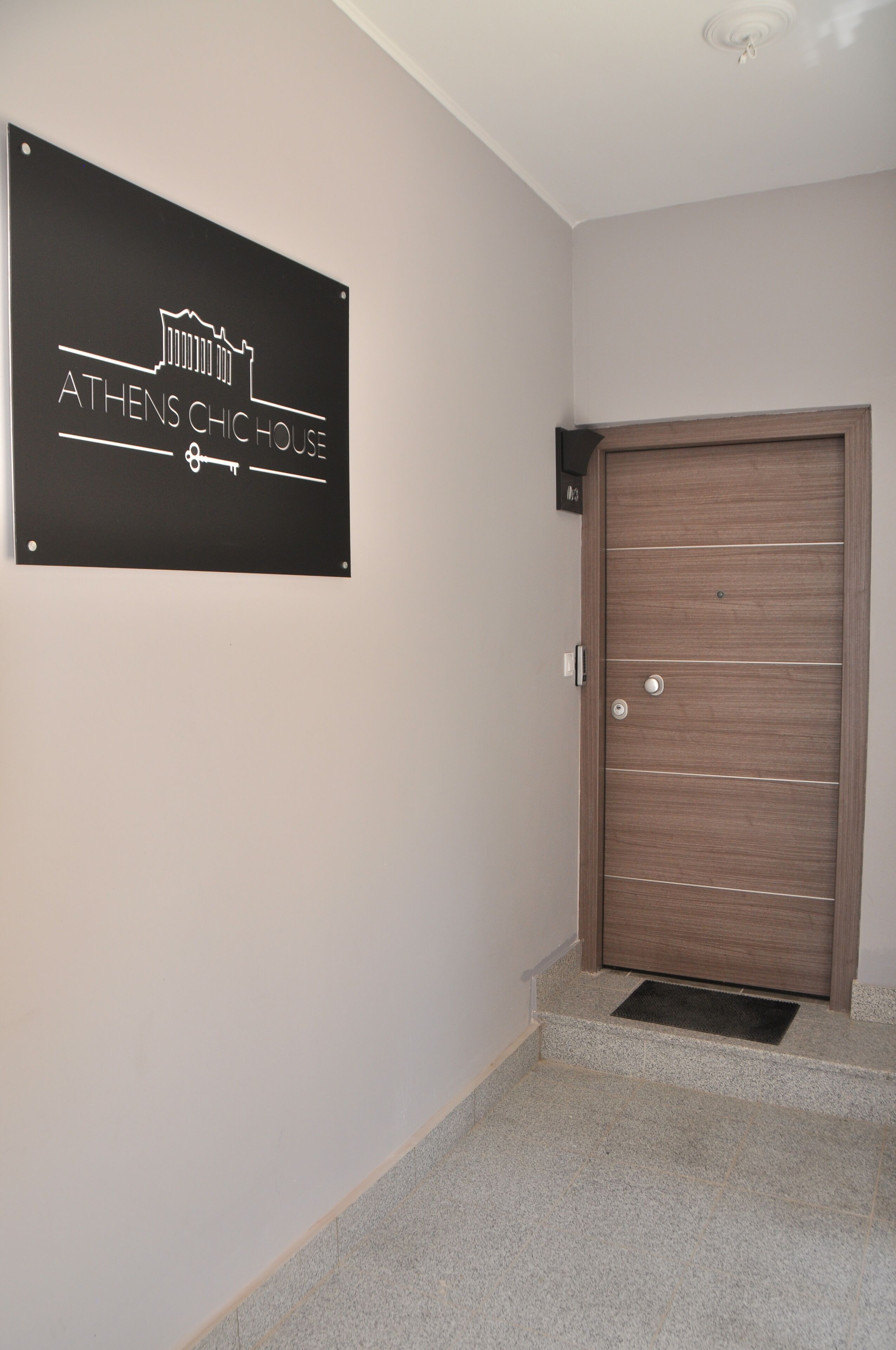 Ein Schild mit der Aufschrift "ATHENS CHIC HOUSE" an einer Wand, eine Holztür und eine Treppe, die zur Tür führt.