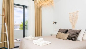 Una cama bien hecha con toallas dobladas, un tapiz de macramé y vistas a través de un ventanal corredero.