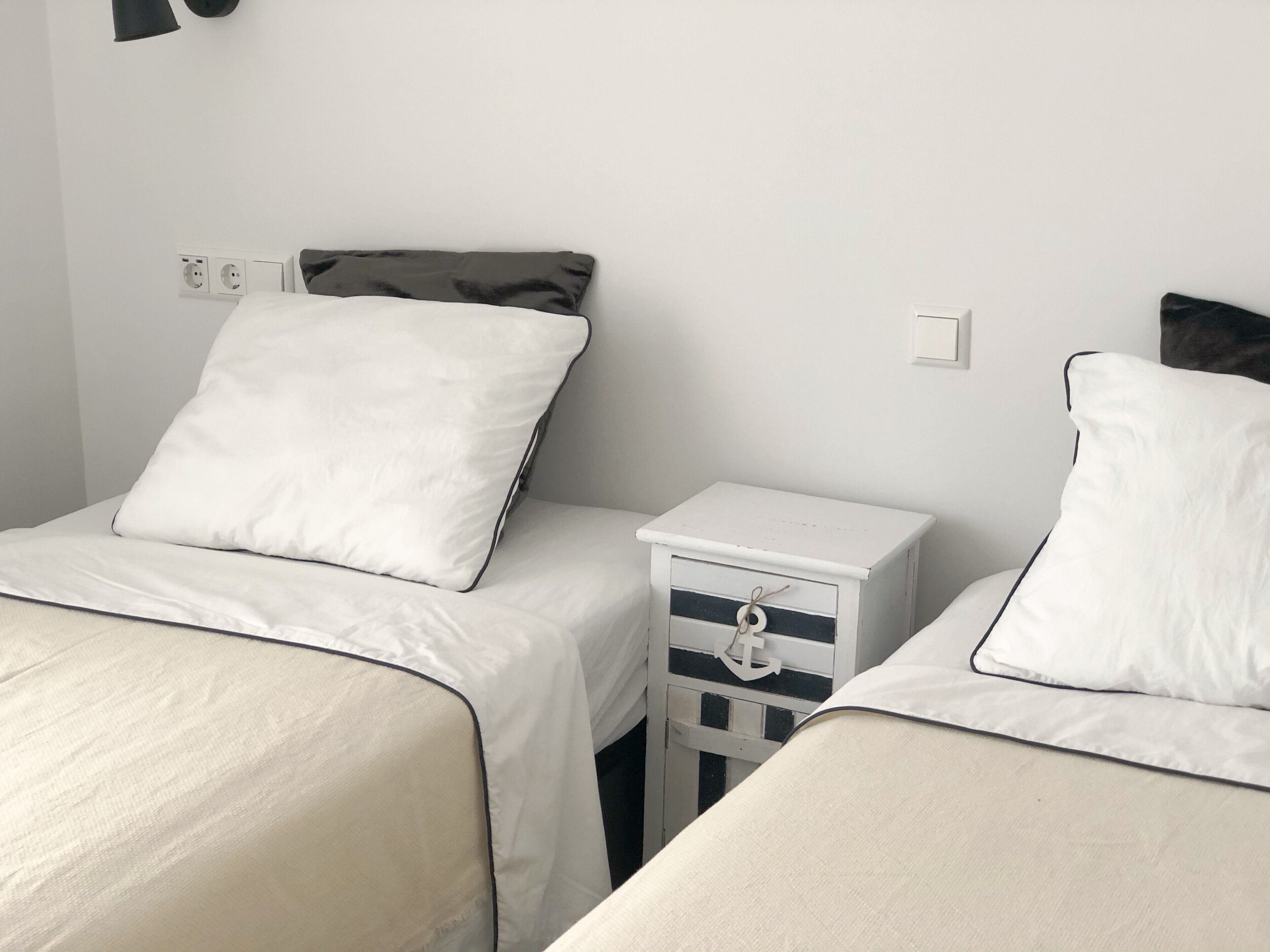 Une chambre avec deux lits, chacun étant équipé de draps blancs et d’un oreiller. On y trouve une petite table de chevet avec un tiroir et une lampe suspendue.