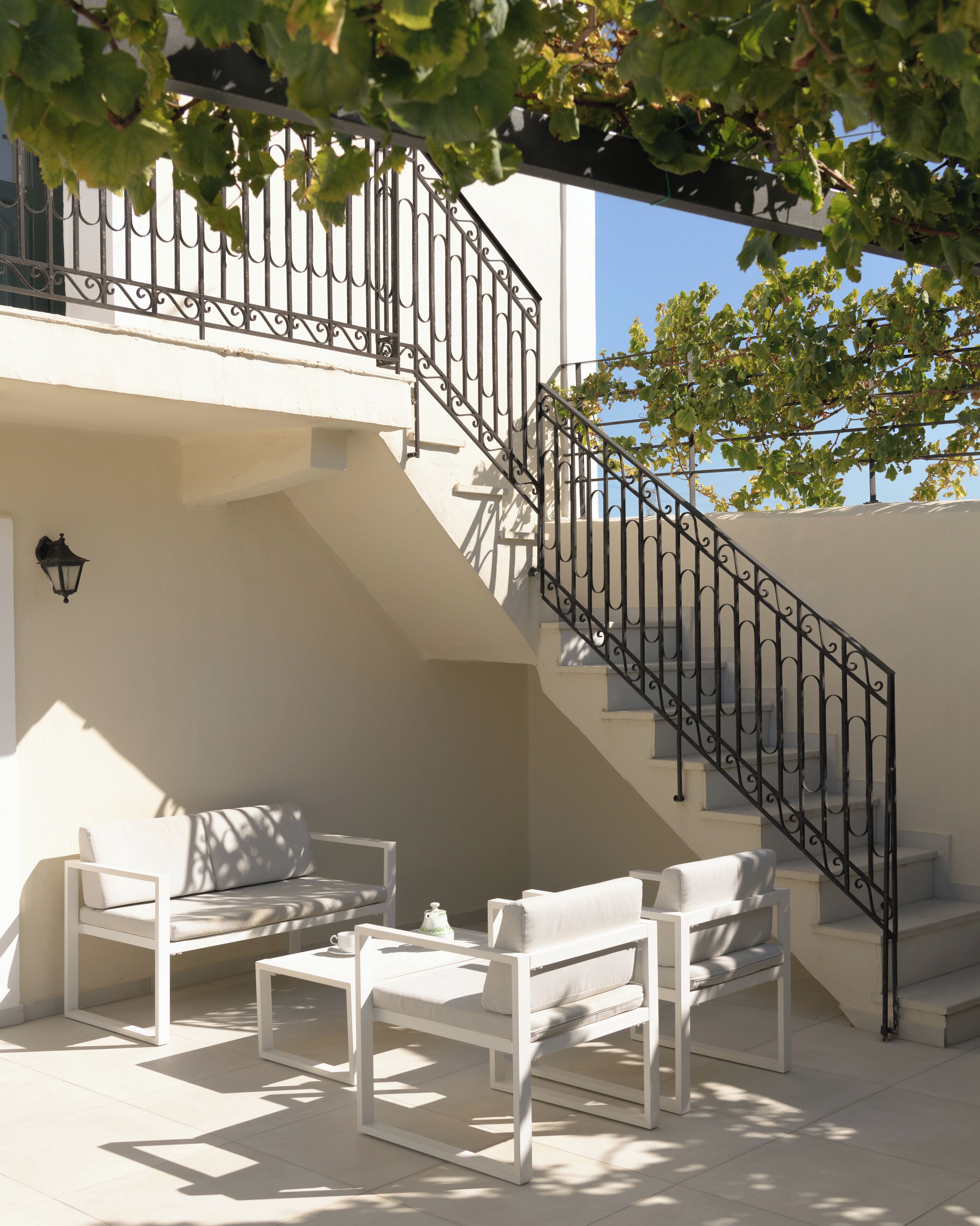 Uma área externa moderna com mobiliário branco, uma escadaria com guarda-corrimão preto e um céu azul claro ao fundo.