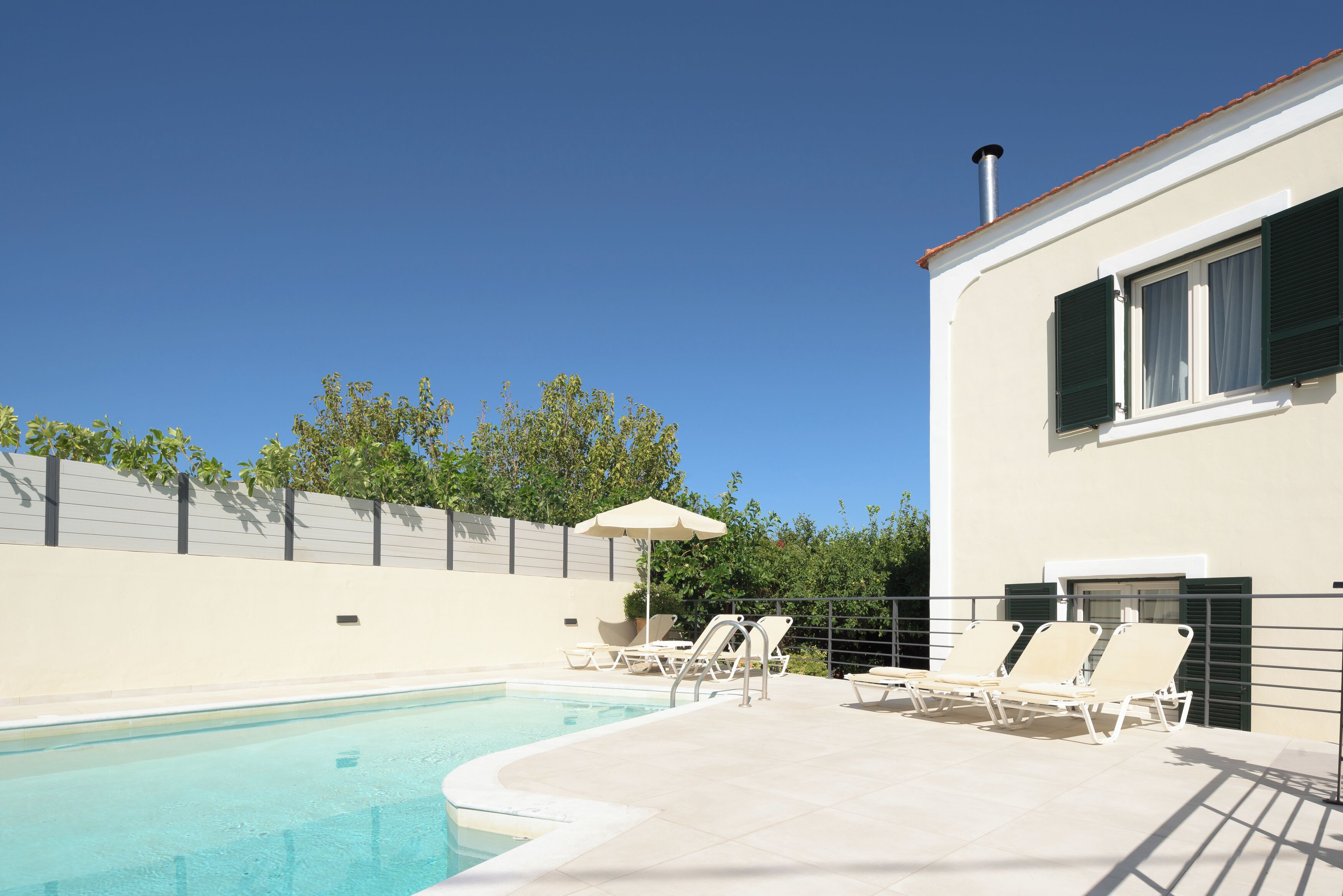 A swimming pool with lounge chairs and an umbrella, surrounded by a low wall and trees.