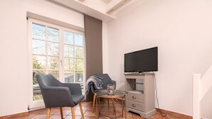 Une pièce avec un téléviseur posé sur un meuble, deux fauteuils gris, une petite table et une fenêtre donnant sur des arbres.