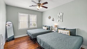 A bedroom with two beds, a ceiling fan, a large window with blinds, and a wooden floor. - M Street Double | Top Floor of a Historic Triplex • Sleeps 12 • Walk to Beach (Galveston)