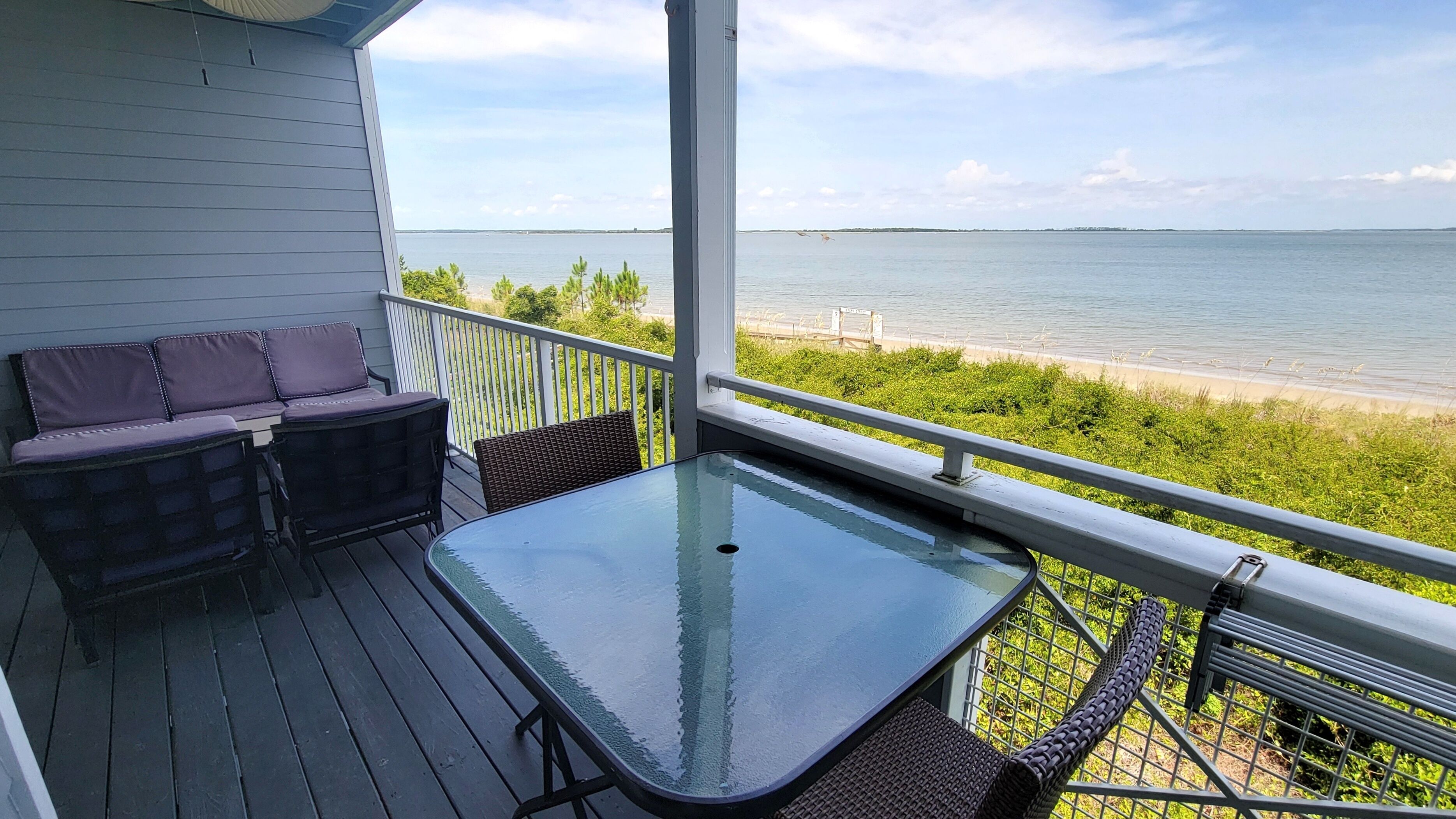 A covered deck with a glass table, two cushioned chairs, and a view of the water.