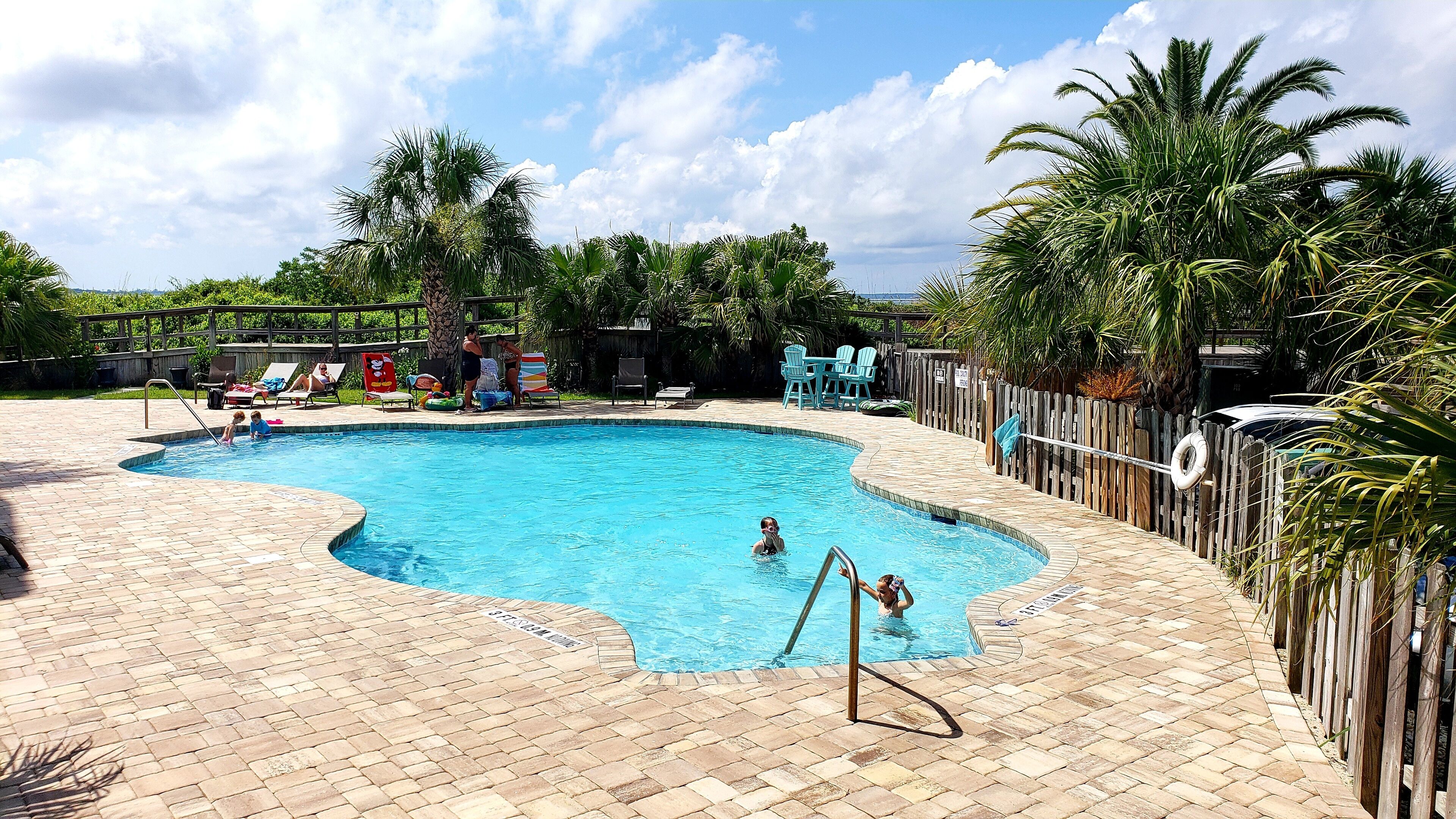 A rectangular swimming pool with a brick deck, surrounded by palm trees and a wooden fence.