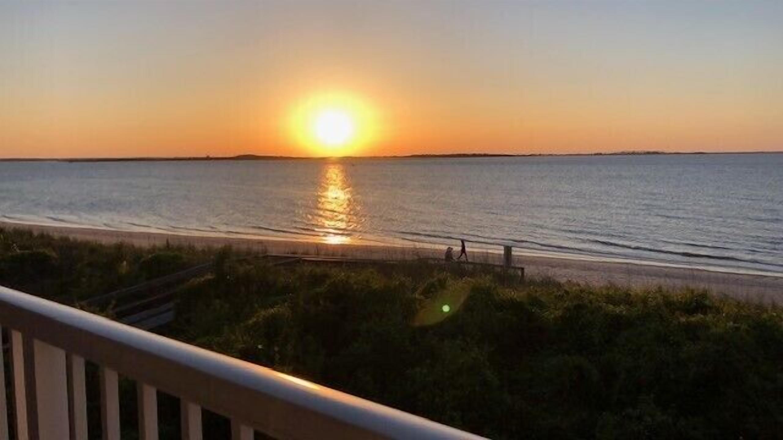 A sunset over a beach with a clear sky, a pathway leading to the water, and a railing in the foreground.