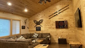 Un intérieur chaleureux de chalet en bois, avec un canapé d’angle, un ventilateur de plafond et un jeu de fléchettes.