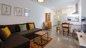 En moderne stue med sofa, sofabord og spiseområde med stole. Et køkken er synligt i baggrunden, med et køleskab og en mikrobølgeovn.