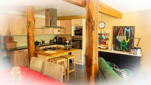 Une cuisine chaleureuse avec des meubles en bois, une cuisinière à gaz et un four moderne. On y trouve un coin repas avec une table et des chaises rouges, ainsi qu’un canapé vert dans le salon.