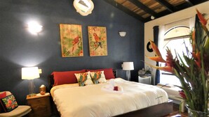 Une chambre avec un grand lit, deux tableaux accrochés au mur, une table de chevet, une lampe, une chaise et un vase rempli de fleurs.