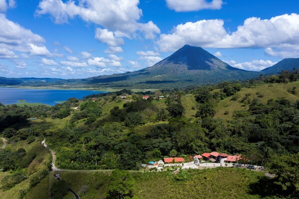 Un paisaje montañoso con un volcán destacado, un lago y un pequeño asentamiento con edificios de techo rojo.