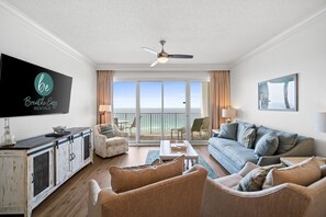 A spacious living room with a large TV, a ceiling fan, a sofa, a coffee table, and a view of the ocean through large windows.