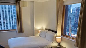 A hotel room with a large bed, a bedside table with a lamp, and a view of tall buildings through the window. - Captivating 2-bed Apartment in Canary Wharf London (London)