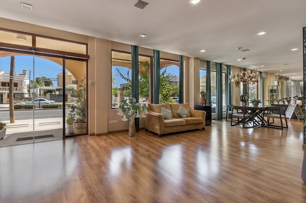 A spacious lobby with a wooden floor, a sofa, a dining table, and large windows offering a view of the street and greenery outside. - Nura Santa Ponsa Pins (Calvia)