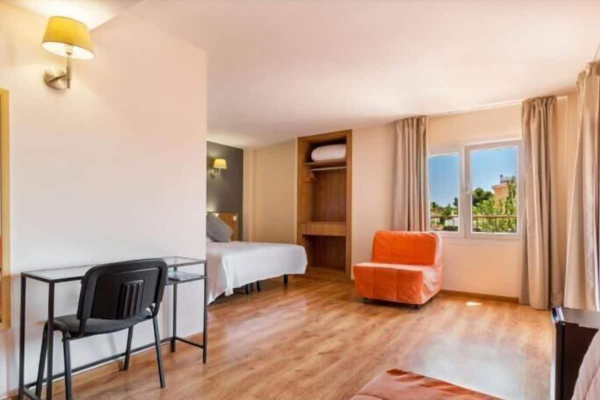 Une chambre d’hôtel comprenant un lit, un bureau avec une chaise, une armoire et une fenêtre avec des rideaux.