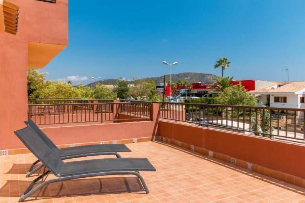 Un balcon avec deux chaises longues, un sol carrelé et une vue sur un paysage urbain avec des bâtiments et une montagne à l’horizon.