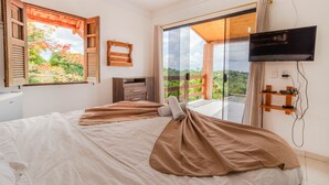 A bedroom with a bed, a TV, a window with wooden shutters, and a view of greenery. - PF12 - Ótima Casa 3 Quartos - Açu da Torre (Mata de São João)