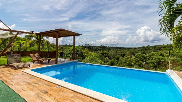 A swimming pool with wooden deck and lounge chairs, surrounded by lush greenery and a clear sky. - PF12 - Ótima Casa 3 Quartos - Açu da Torre (Mata de São João)